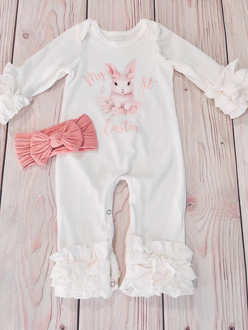 K&ouml;nnte beinhalten: Wei&szlig;er Baby-Strampler mit einem rosa Hasen-Motiv und dem Text "My 1st Easter". Der Strampler hat R&uuml;schen an den &Auml;rmeln und Beinen. Ein rosa Stirnband ist im Lieferumfang enthalten.