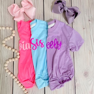 Może przedstawiać: Trzy body z kr&oacute;tkim rękawem w kolorze r&oacute;żowym, jasnoniebieskim i lawendowym, każde z napisem kursywą. R&oacute;żowe body ma napis 'via', niebieskie 'Sis', a lawendowe 'eely'. Dołączono dwie duże kokardy do włos&oacute;w w kolorze jasnor&oacute;żowym i lawendowym.