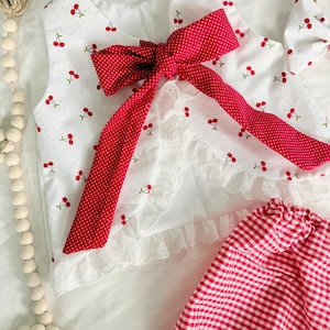 May include: A white sleeveless top featuring a red cherry print, a large red polka-dot bow at the neckline, and white eyelet lace trim along the hem. The set includes red and white gingham shorts and a matching hair accessory with a cherry print.