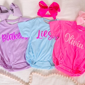 May include: Three short-sleeve rompers in lavender, light blue, and hot pink. The lavender romper features the name Blakeley in pink, the light blue one says Lil Sis in pink, and the hot pink one says Olivia in white. Each has a matching hair bow.