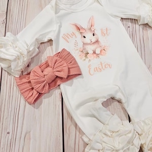 K&ouml;nnte beinhalten: Wei&szlig;er Baby-Strampler mit einem rosa Hasen-Motiv und dem Text "My 1st Easter". Der Strampler hat R&uuml;schen&auml;rmel und einen R&uuml;schen-Saum. Ein rosa Schleifen-Stirnband ist im Lieferumfang enthalten.