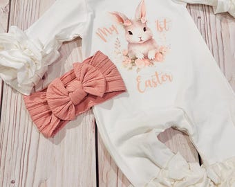 May include: White baby onesie with a pink bunny rabbit graphic and the text "My 1st Easter". The onesie has ruffled sleeves and a ruffled bottom. A pink bow headband is included.