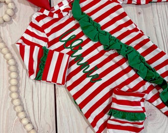May include: A red and white striped outfit with green ruffle accents and the name "Winnie" embroidered in green. A large red bow is at the top. The outfit is on a light wood surface.