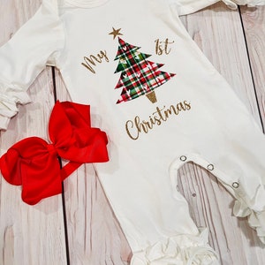 May include: White baby onesie with a green, red, and white plaid Christmas tree with gold glitter and the text "My 1st Christmas". A red bow is included.