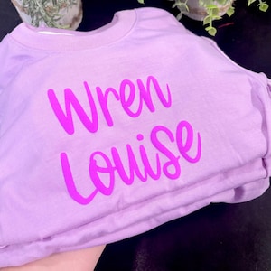 Może przedstawiać: Jasnofioletowa bluza z okrągłym dekoltem i napisem "Wren Louise" nadrukowanym pogrubioną, jaskrawor&oacute;żową czcionką skryptową na środku klatki piersiowej. Odzież posiada prążkowany dekolt oraz długie rękawy i jest zaprezentowana w złożonym stosie.