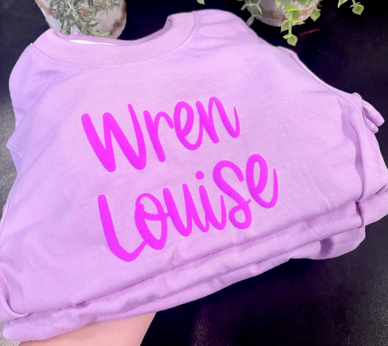 Może przedstawiać: Jasnofioletowa bluza z okrągłym dekoltem i napisem "Wren Louise" nadrukowanym pogrubioną, jaskrawor&oacute;żową czcionką skryptową na środku klatki piersiowej. Odzież posiada prążkowany dekolt oraz długie rękawy i jest zaprezentowana w złożonym stosie.