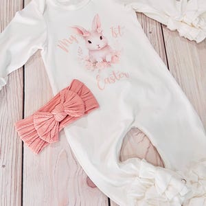 K&ouml;nnte beinhalten: Wei&szlig;er lang&auml;rmliger Baby-Strampler mit einem rosa Hasen-Motiv und dem Text "My 1st Easter". Der Strampler hat R&uuml;schen an den &Auml;rmeln und Beinen. Ein rosa Schleifen-Stirnband ist im Lieferumfang enthalten.