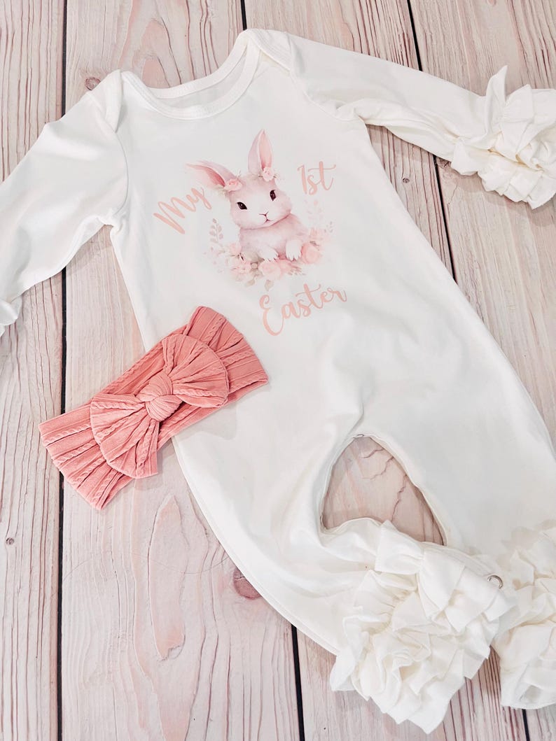 K&ouml;nnte beinhalten: Wei&szlig;er lang&auml;rmliger Baby-Strampler mit einem rosa Hasen-Motiv und dem Text "My 1st Easter". Der Strampler hat R&uuml;schen an den &Auml;rmeln und Beinen. Ein rosa Schleifen-Stirnband ist im Lieferumfang enthalten.