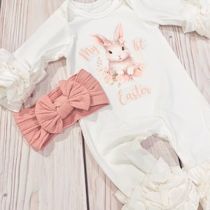 K&ouml;nnte beinhalten: Wei&szlig;er Baby-Strampler mit einem rosa Hasen-Motiv und dem Text "My 1st Easter". Der Strampler hat wei&szlig;e R&uuml;schen an den &Auml;rmeln und Beinen. Ein rosa Schleifen-Stirnband ist im Lieferumfang enthalten.