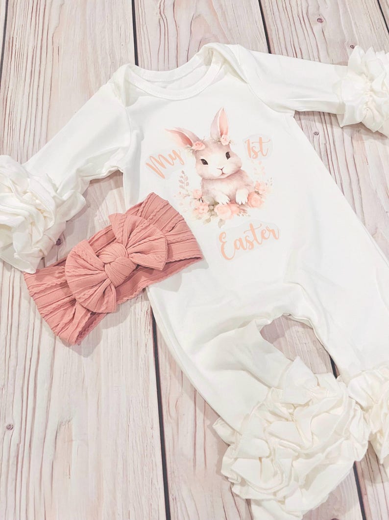 K&ouml;nnte beinhalten: Wei&szlig;er Baby-Strampler mit einem rosa Hasen-Motiv und dem Text "My 1st Easter". Der Strampler hat wei&szlig;e R&uuml;schen an den &Auml;rmeln und Beinen. Ein rosa Schleifen-Stirnband ist im Lieferumfang enthalten.