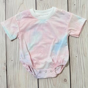Może przedstawiać: Body niemowlęce z kr&oacute;tkim rękawem z wzorem tie-dye w delikatnych pastelowych odcieniach r&oacute;żu, błękitu i bieli. Ubranko posiada jednolity biały okrągły dekolt oraz trzy metalowe zatrzaski w kroku, kt&oacute;re zapewniają bezpieczne i łatwe zapięcie.