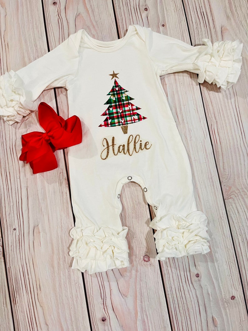 May include: White baby onesie with a green, red, and white plaid Christmas tree design and the name "Hallie" in gold glitter lettering. The onesie has white ruffles on the sleeves and legs.