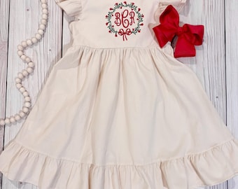 Könnte beinhalten: Ein cremefarbenes Kleid mit gerüschten Ärmeln und einem gerüschten Saum. Das Oberteil zeigt einen gestickten grünen Kranz mit roten Beeren und einer roten Schleife, der die Monogrammbuchstaben BCR in Rot umgibt. Eine große rote Haarschleife liegt daneben.