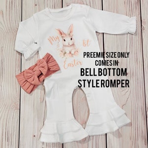 K&ouml;nnte beinhalten: Wei&szlig;er Baby Strampler mit einem Hasen- und Blumendesign. Der Strampler sagt "Mein 1. Ostern". Er kommt im Glockenbein-Stil. Ein pfirsichfarbenes Stirnband ist enthalten.