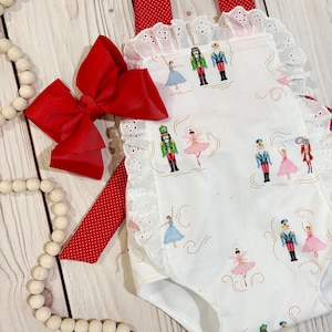 May include: A white romper featuring a repeating pattern of nutcrackers and ballerinas, accented with red and white polka dot straps and white eyelet lace trim. A large red bow is attached to the side, complementing the festive holiday design.