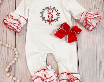 May include: A white long-sleeve romper featuring ruffled cuffs and leg openings with red trim. The chest displays an embroidered green wreath with a red letter S and a red bow. A large red hair bow with white scalloped edges is placed on the romper.