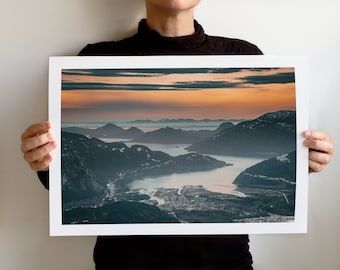 May include: A panoramic view of a mountain range with a valley and a body of water in the foreground. The sky is a vibrant orange and pink, suggesting a sunset or sunrise. The mountains are covered in trees and appear to be forested.