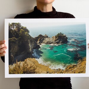 May include: A panoramic view of a rocky coastline with a turquoise blue ocean. The water is clear and calm, and the rocks are covered in lush green vegetation. A small waterfall cascades down the cliff face, creating a picturesque scene.