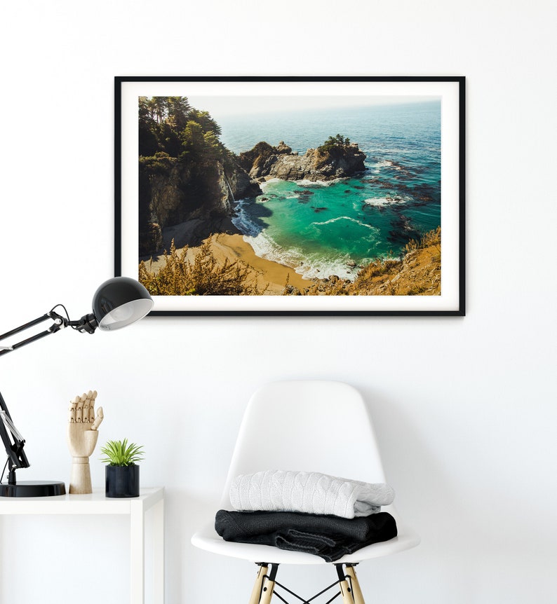 May include: A framed photograph of a scenic coastal landscape. The image shows a rocky cove with a small beach and turquoise water. Lush green trees and cliffs surround the cove. The water is clear and calm, with a few small waves breaking on the shore.