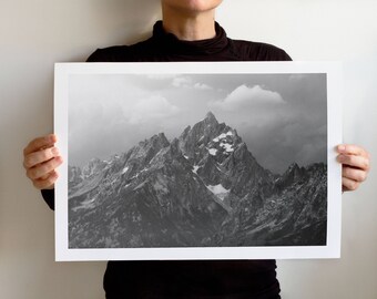 May include: Black and white print of a mountain range with snow-capped peaks and cloudy skies. The print is held by a person and has a white border. The image captures the rugged beauty of the mountains.