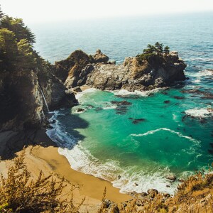 May include: A view of a secluded cove with a waterfall cascading down a rocky cliff. The water is a vibrant turquoise blue, and the beach is a soft, golden sand. Lush green trees line the cliffside, creating a picturesque scene.