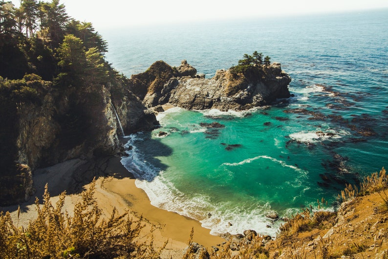 May include: A view of a secluded cove with a waterfall cascading down a rocky cliff. The water is a vibrant turquoise blue, and the beach is a soft, golden sand. Lush green trees line the cliffside, creating a picturesque scene.