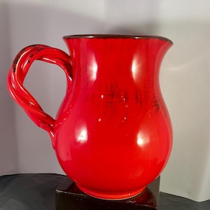 May include: A bright red ceramic jug with a twisted handle. The jug has a glossy finish and a dark brown rim. The handle features a unique, rope-like design. The jug is displayed on a dark wooden base.