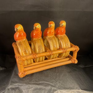 Può includere: Un set di quattro portatovaglioli a forma di pappagallo in un supporto di bambù intrecciato. I pappagalli hanno teste arancioni e gialle con corpi marroni. Il supporto in bambù è di colore marrone chiaro.