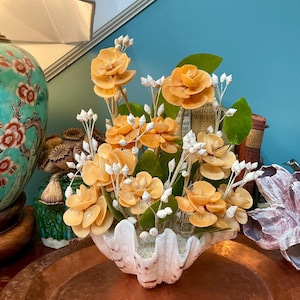May include: A decorative arrangement featuring peach-coloured flowers crafted from seashells, accented with small white spiral shells and green leaves. The floral display is set within a large, white, fluted clam shell base filled with sand.