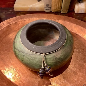 Puede incluir: Un cuenco de madera redondeado de color verde con un borde gris oscuro y un acabado liso y pulido. Un cordón fino de color claro está atado alrededor del cuello del cuenco, con dos pequeñas cuentas de vidrio esféricas y multicolores.