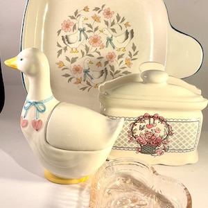 May include: A selection of cream-coloured ceramic kitchen items, including a duck-shaped container, a heart-shaped dish, a lidded container, and a serving dish. The pieces feature floral and duck designs, with pink, blue, and yellow accents.