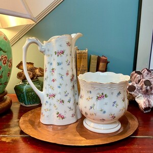 May include: A white porcelain jug and a matching bowl, both decorated with a delicate pattern of small pink, blue and yellow flowers. The jug features a curved handle and a scalloped rim, while the bowl has a wide, fluted opening.