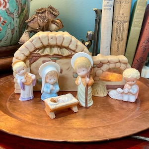 May include: A ceramic nativity set featuring a stable backdrop and five small figurines including an angel with a harp, a figure in blue, a central figure with a staff, a baby in a manger, and a kneeling figure with a lamb, all on a copper tray.