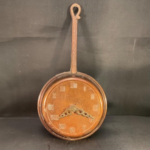 Peut inclure: Une horloge murale de couleur cuivrée avec une poignée. Le cadran de l'horloge comporte des chiffres romains et des aiguilles décoratives. La poignée est longue et comporte une boucle en haut. L'horloge est ronde et a une texture martelée.