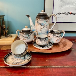 May include: A porcelain tea set featuring a teapot, sugar bowl, creamer, and two teacups with saucers. Each piece is decorated with a raised dragon motif in black, white, and blue, accented with gold trim on a white and dark grey gradient background.