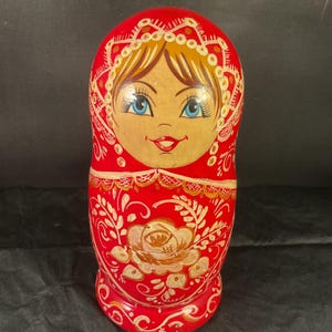 May include: A red Matryoshka doll with a painted face and floral design. The doll features a smiling face with blue eyes, blonde hair, and a red headscarf. The body is adorned with a gold and white floral pattern.