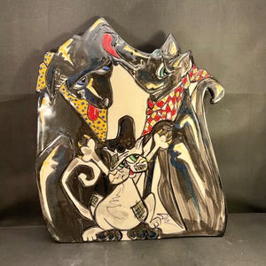 May include: A decorative ceramic wall plaque featuring a hand-painted illustration of two black dogs and a white cat. The dogs wear colorful patterned bandanas, and the cat is positioned in the center. The artwork uses bold black outlines.