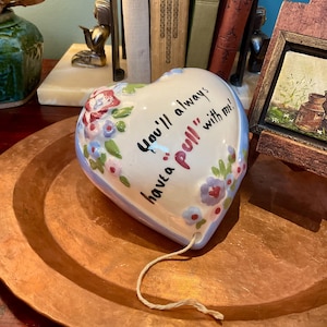 May include: A heart-shaped ceramic wall plaque with a glossy white finish, featuring hand-painted blue, pink, and green floral accents. The text "You'll always have a pull with me!" is printed in black and red, with a string extending from the bottom.