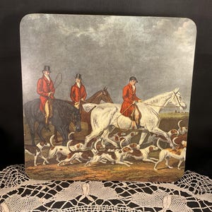 Puede incluir: Un posavasos decorativo cuadrado con una escena de caza. Tres jinetes con abrigos rojos a caballo lideran una jauría de perros por un campo. El fondo es un cielo nublado.