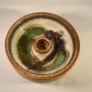 Può includere: Un portacandele in ceramica fatto a mano con un design circolare. Il portacandele presenta un pozzetto centrale ed è decorato con sfumature di marrone, verde, blu e bianco. Il bordo ha una fascia blu scuro.