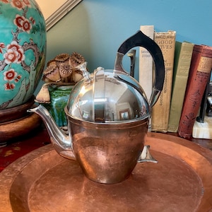 Op de afbeelding: Een gepolijste zilverkleurige metalen theepot met een rond lichaam, een gebogen tuit en een zwart boogvormig handvat. De theepot heeft een koepelvormig deksel met een kleine knop en een metalen afwerking die de omgeving reflecteert.