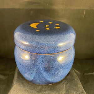 Könnte beinhalten: Eine blaue Keramikdose mit Deckel. Der Deckel ist mit einem goldenen Mond und Sternen verziert. Die Dose hat eine gesprenkelte Textur und eine runde Form. Die Dose steht auf einer dunklen Oberfläche.