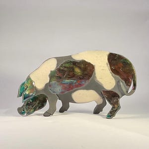 Könnte beinhalten: Eine Keramikschwein-Skulptur mit grauem Körper, cremefarbenen Flecken und farbenfrohen Akzenten. Das Schwein wird in einer Futtersuchhaltung dargestellt, mit der Schnauze am Boden. Die Skulptur hat eine glänzende Oberfläche und steht vor einem weißen Hintergrund.