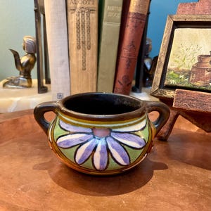May include: A small, round ceramic bowl with two side handles, featuring a hand-painted floral motif with purple petals, green leaves, and a brown rim. The bowl has a visible crackled glaze finish and a dark brown interior, showcasing a decorative design.