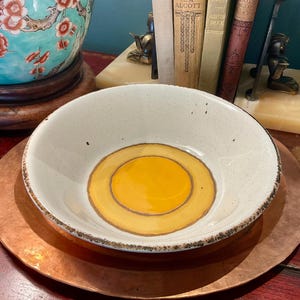 Peut inclure: Un bol en céramique rond avec un émail blanc cassé moucheté et un bord brun foncé. L'intérieur présente un centre jaune moutarde lisse et circulaire, entouré d'un anneau ocre légèrement plus foncé, formant un motif de cible distinct.