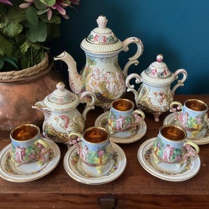 Puede incluir: Un juego de té de porcelana vintage con una tetera grande, un azucarero, una crema y seis tazas de té con platillos. El juego está decorado con figuras clásicas en una paleta de colores pastel, con ribetes dorados.
