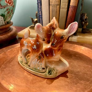 Peut inclure: Une figurine en céramique représentant un faon debout et un cerf couché. Les cerfs sont peints dans des tons marron et crème avec des taches blanches sur le faon. Ils sont placés sur une petite base ovale avec des touches de vert représentant l'herbe.