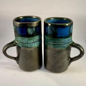 May include: Two ceramic mugs with a dark brown base and a gradient of teal and blue at the top. The mugs have a glossy finish and a handle on the side. The interior of the mugs is a deep blue colour.