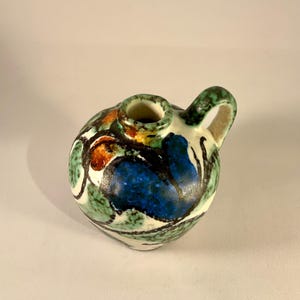 Può includere: Una piccola brocca rotonda in ceramica con manico. La brocca è dipinta con un design colorato con forme astratte blu, verdi, arancioni e nere. Il bordo e il manico sono verdi.