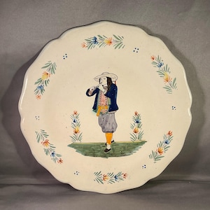 Könnte beinhalten: Ein weißer Keramikteller mit gewelltem Rand, der eine handgemalte Illustration einer Person zeigt, die Flöte spielt. Die Person ist in historischer Kleidung gekleidet. Blumenmotive in Blau, Orange und Grün verzieren den Rand.